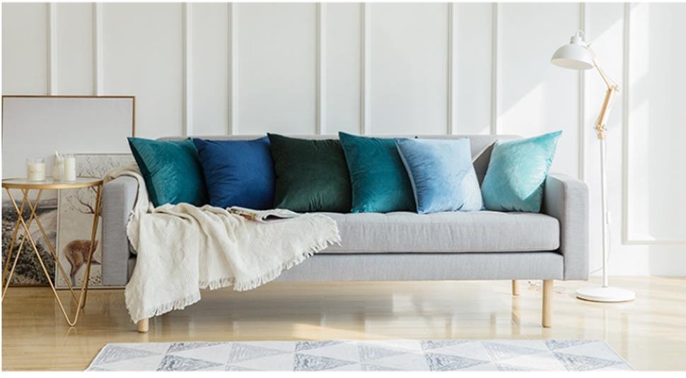 Assortment of velvet throw pillow covers in various shades of blue and green arranged on a gray sofa with a white blanket.