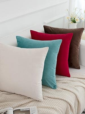 Four velvet throw pillow covers in cream, teal, red, and brown arranged on a couch with a blanket.