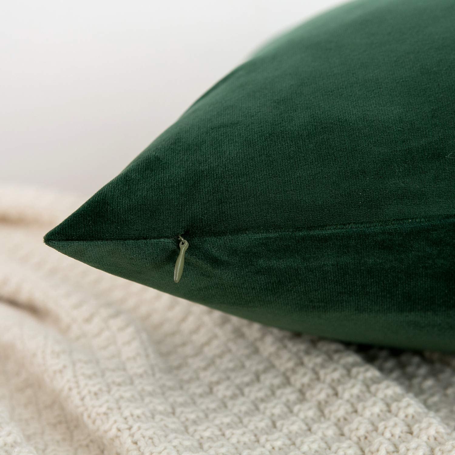 Close-up of a dark green velvet throw pillow cover with a visible zipper, resting on a cream-colored knitted blanket.