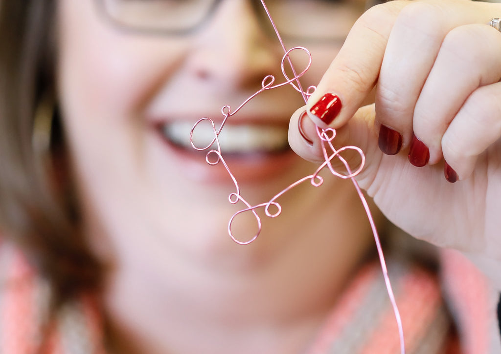 Close-up of hands shaping pink wire into a heart shape, demonstrating jewelry making.