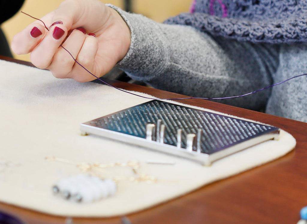 Close-up of a person's hands holding purple wire over a metal jig board with pegs for jewelry making.