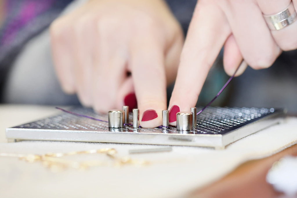 Close-up of hands using a metal wire jig with pegs to create jewelry, with purple wire being manipulated.
