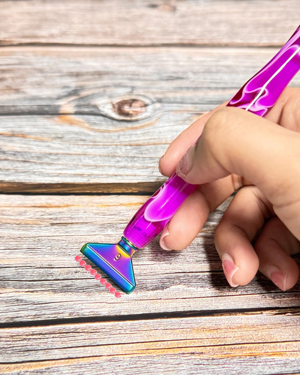 Hand holding a purple diamond painting pen with a multi-colored metal tip applying pink beads to a wooden surface.