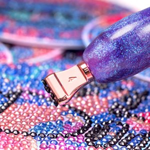 Diamond painting pen with rose gold tip placing a pink gem on a colorful canvas
