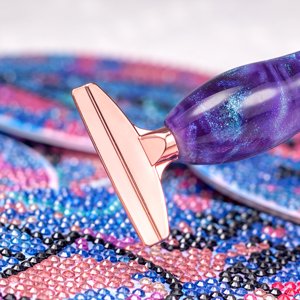 Close-up of a galaxy purple rose gold diamond painting pen with a rose gold tip resting on a colorful diamond art canvas.