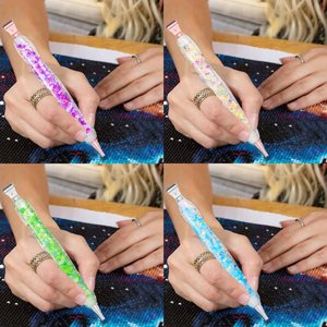 Four diamond painting pens with glitter in purple, yellow, green, and blue, held by hands working on a diamond painting canvas.