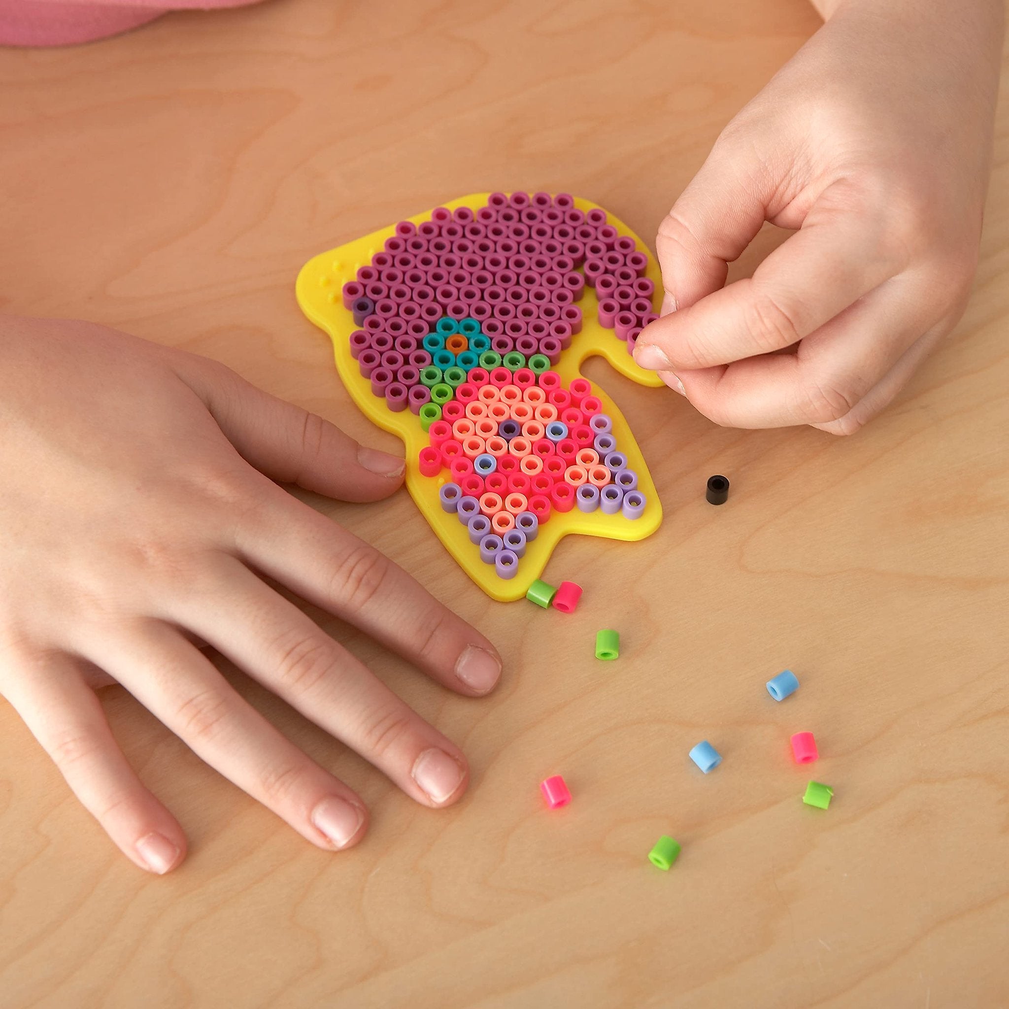 Child's hands placing colorful fuse beads onto a yellow pegboard to create a dinosaur shape.