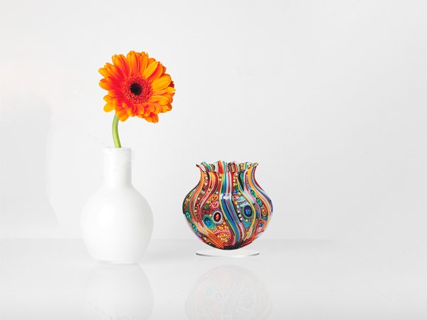 Colorful 5D diamond painting vase ornament next to a white vase with an orange gerbera daisy on a reflective surface.