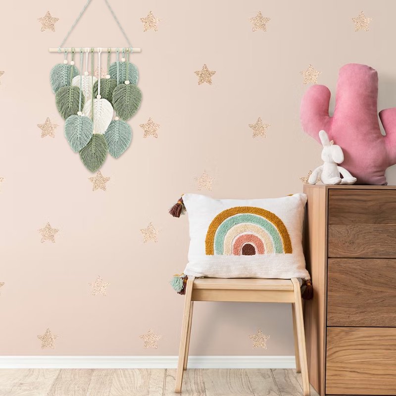 Macrame leaf wall hanging with wooden beads and stick, displayed in a nursery with a rainbow pillow and cactus plush.