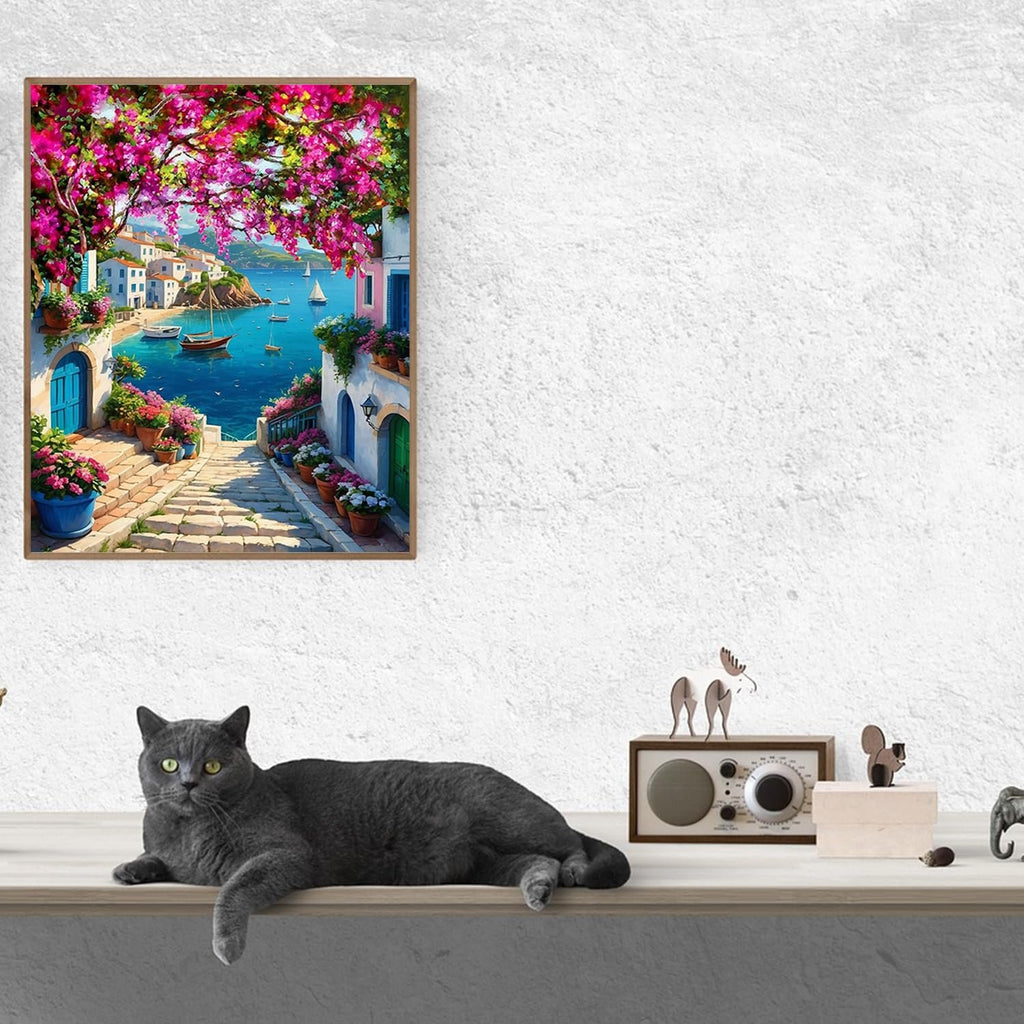 Paint by numbers canvas depicting a Mediterranean village scene with a blue sea and pink flowers, framed and hung on a wall above a shelf with a cat and decorative items.