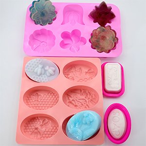 Two silicone soap molds and two soap bars, one pink oval mold with a bar of soap, one pink rectangular mold with a bar of soap, one pink mold with bee and honeycomb designs, one pink mold with floral designs, and one pink mold with floral designs.