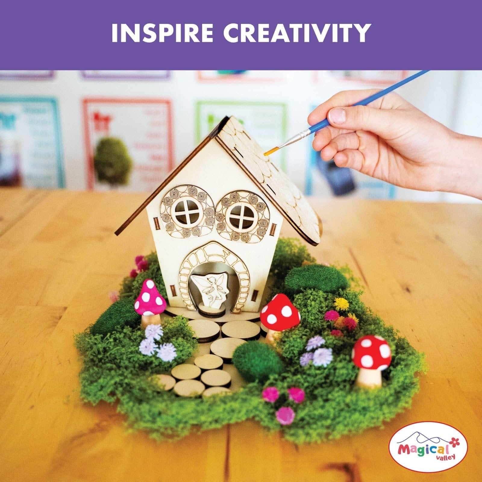 Child painting a wooden fairy house craft kit with moss, flowers, and mushrooms on a wooden table.