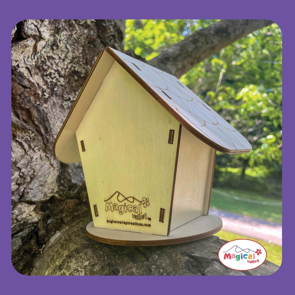 Wooden fairy house craft kit on a tree branch, showing the assembled house with engraved details and a logo.