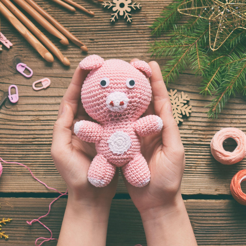 Hands holding a pink crocheted pig toy with googly eyes and a white belly, surrounded by crochet hooks, yarn, and festive decorations on a wooden background.