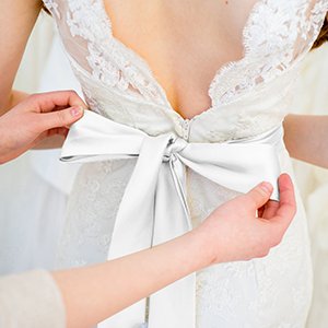 Close-up of hands tying a white satin ribbon bow on the back of a wedding dress.