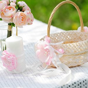 Wedding decor setup with a white candle, pink flowers, and a decorative basket on a white tablecloth outdoors.