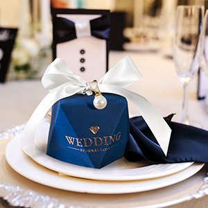 Blue geometric wedding favor box tied with white satin ribbon and a pearl charm, set on a plate with a navy napkin.