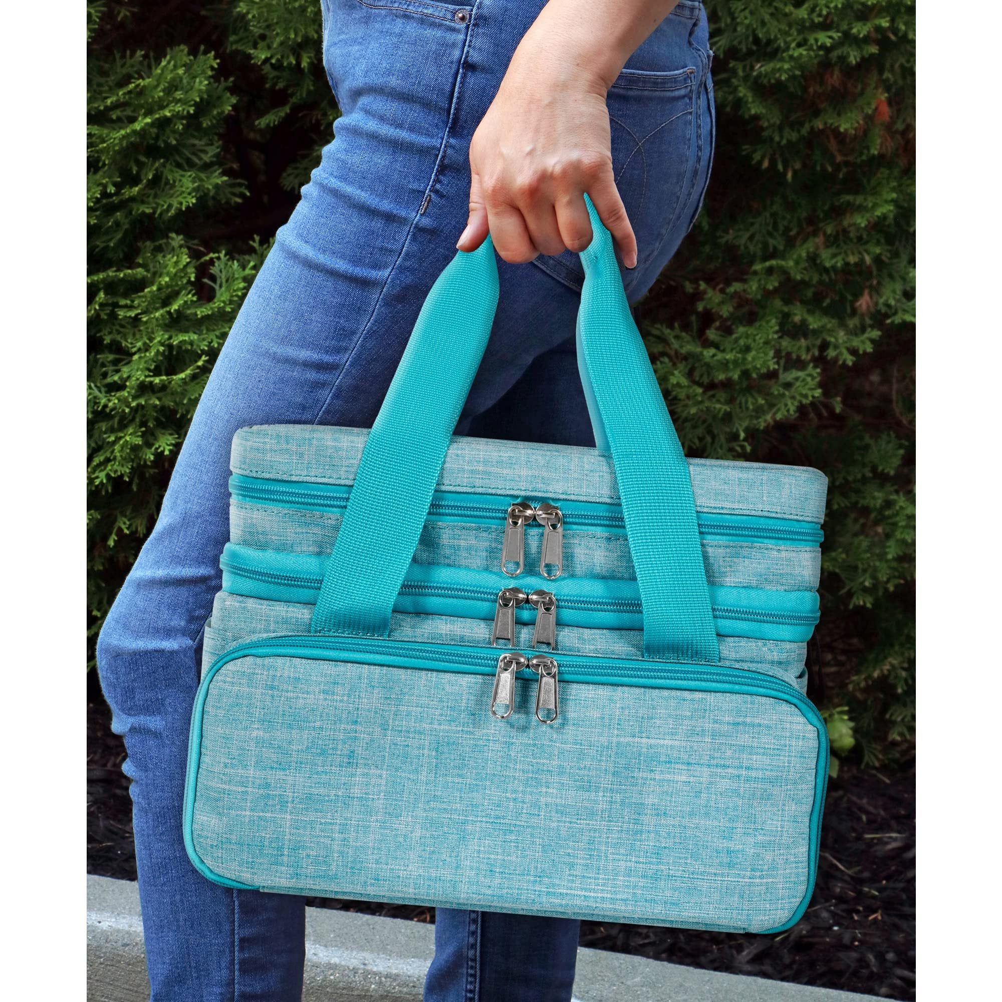 Person in jeans holding a teal double-layer sewing storage bag with multiple zippers outdoors.
