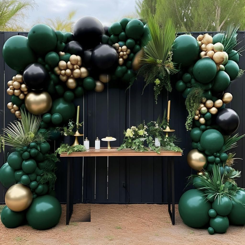 Dark green, black, and metallic gold balloon garland arching over a wooden table with greenery and candles, set against a black fence.