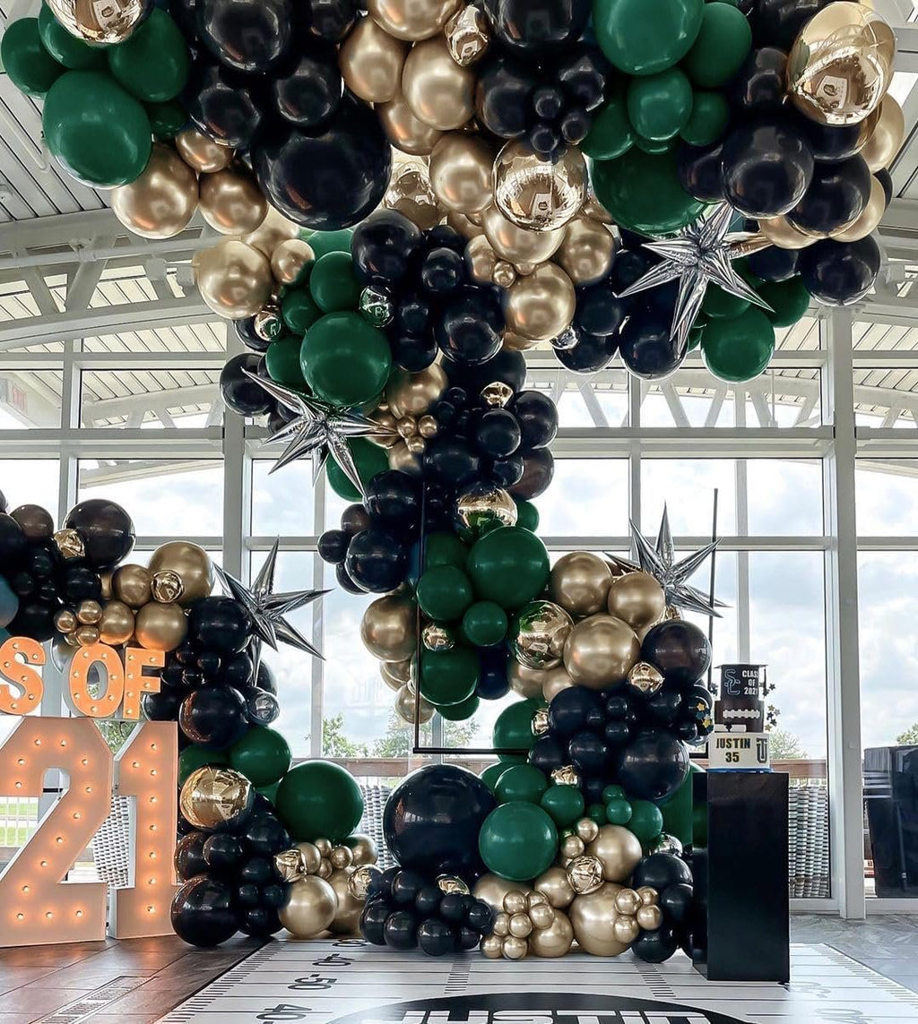 Dark green, black, and metallic gold balloon garland arch with star accents, set up for a graduation party with large light-up numbers and a cake stand.