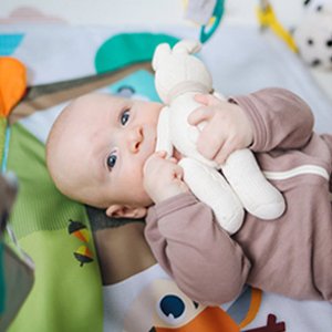 Baby lying on a playmat holding a stuffed bunny toy, looking at the camera.
