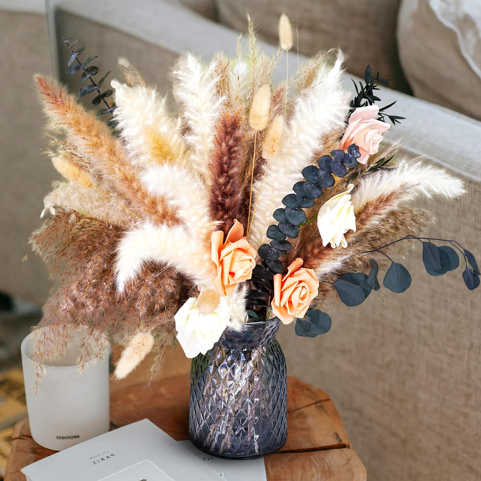 Dried flower bouquet with pampas grass and roses in a textured vase on a wooden table next to a candle and book.