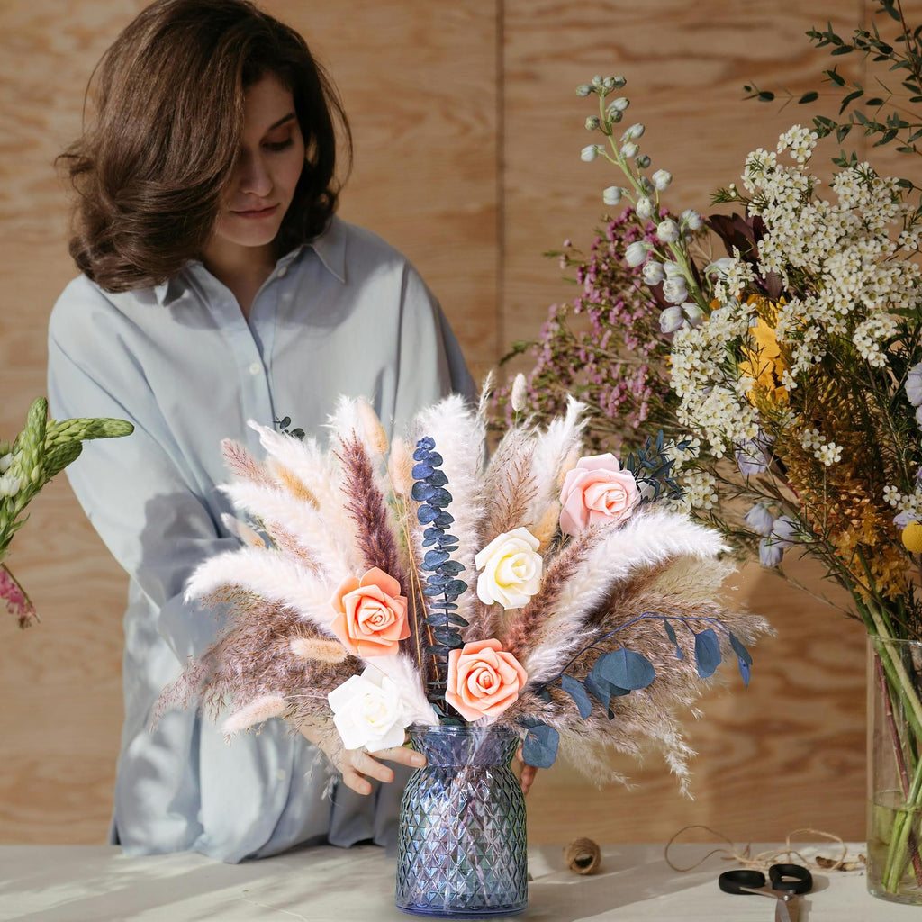 Woman arranging a dried flower bouquet with pampas grass and roses in a blue glass vase.