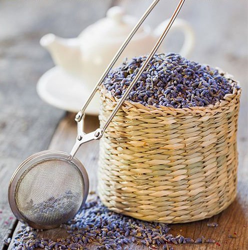 Woven basket filled with dried lavender buds next to a tea infuser on a wooden surface, with a teapot in the background.