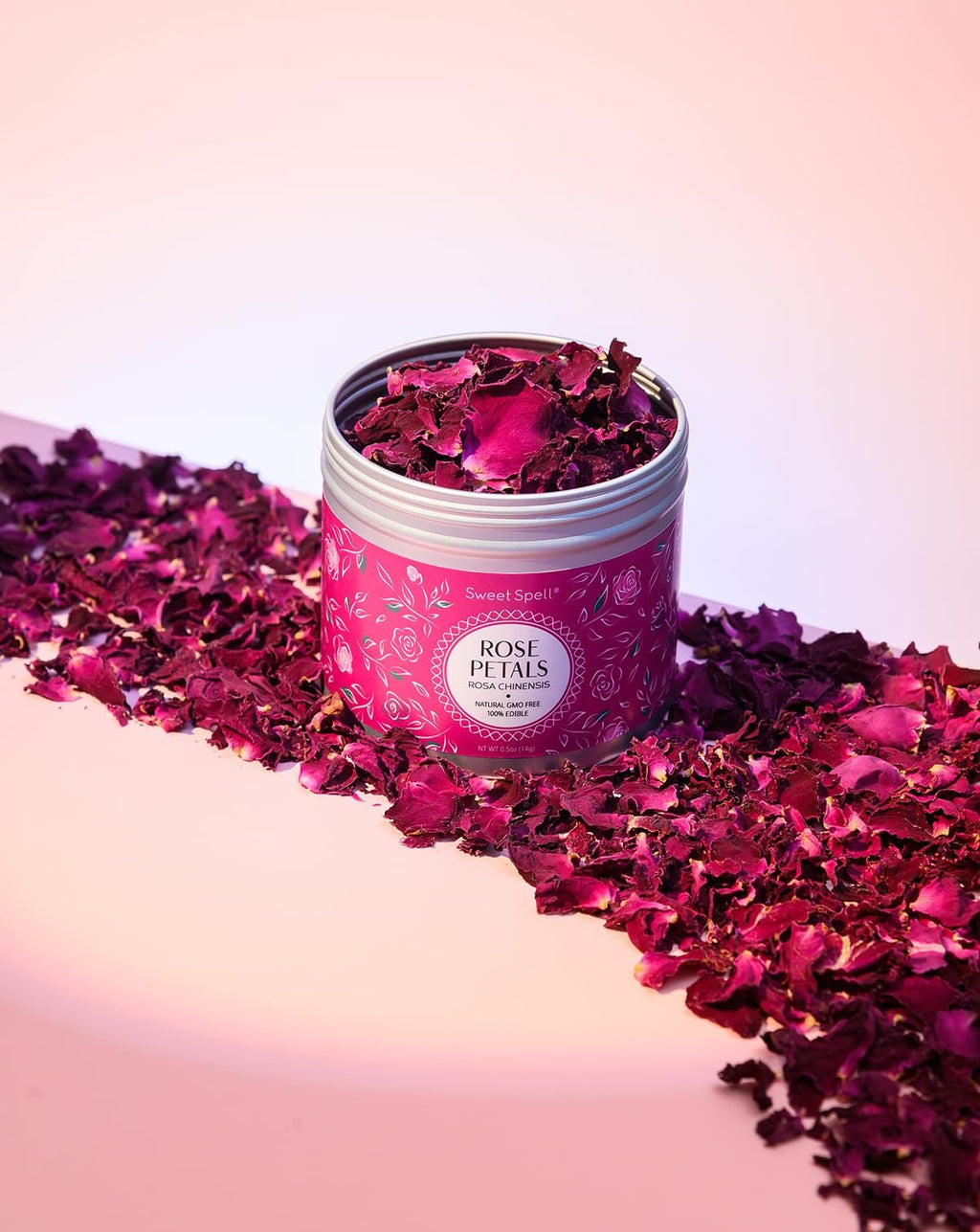 Can of dried rose petals with loose petals scattered around, on a pink and white surface.