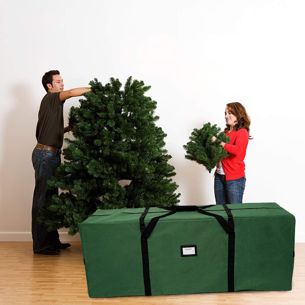 Man and woman assembling a large artificial Christmas tree next to a green storage bag.
