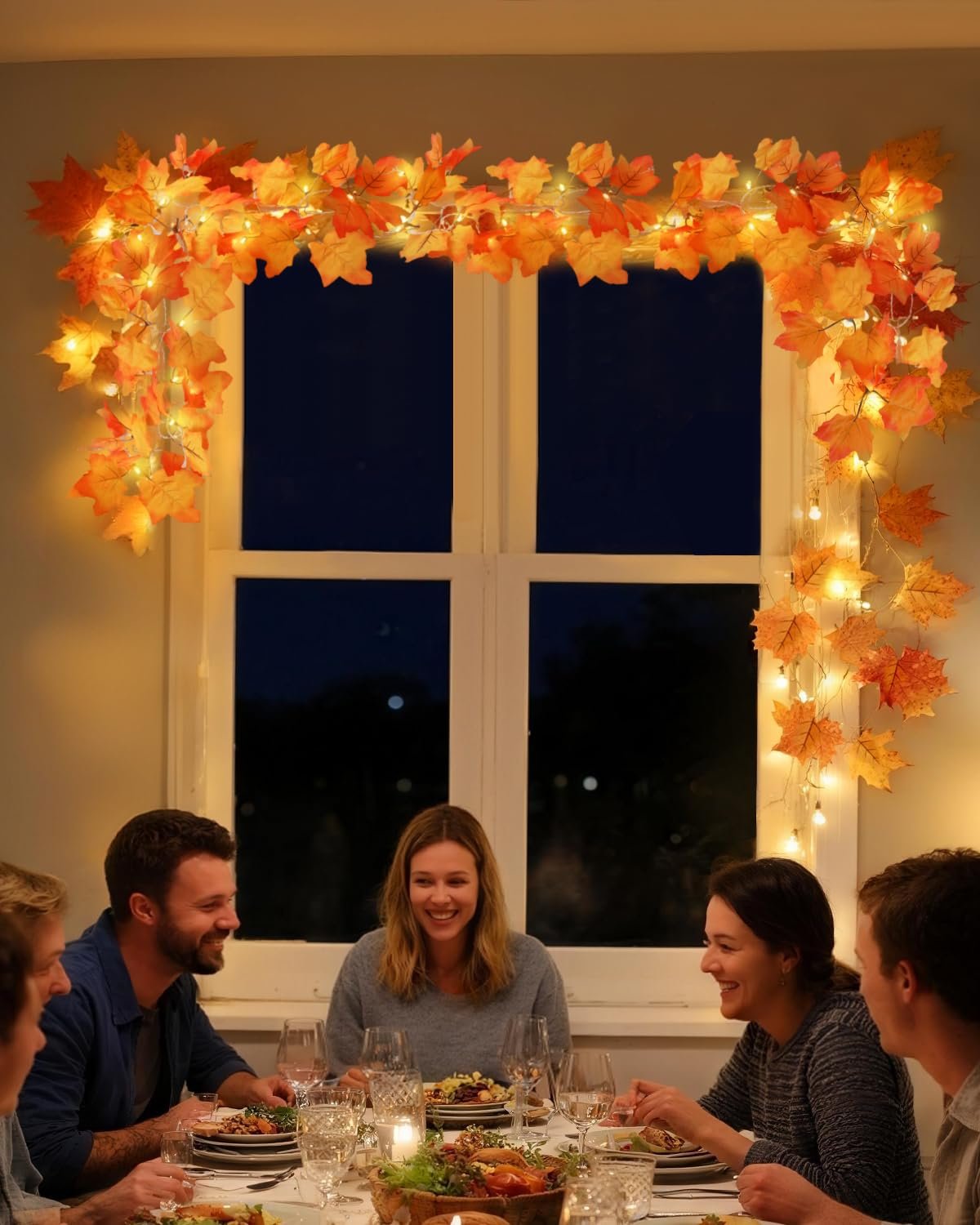 Family and friends enjoying a festive dinner with autumn leaf garland and warm LED lights above a window.