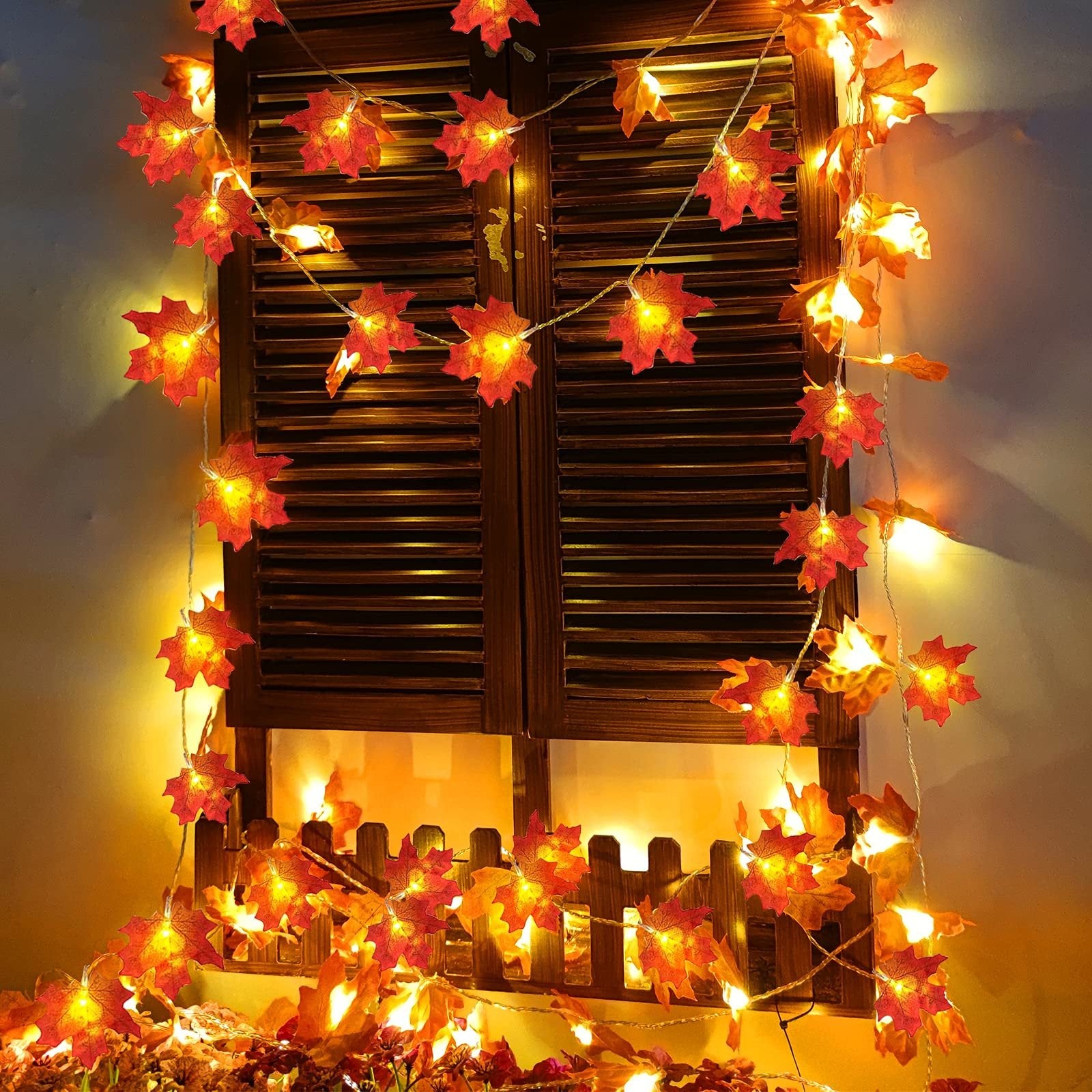 Fall maple leaf LED string lights draped across a wooden shutter and fence, illuminated with warm yellow light.