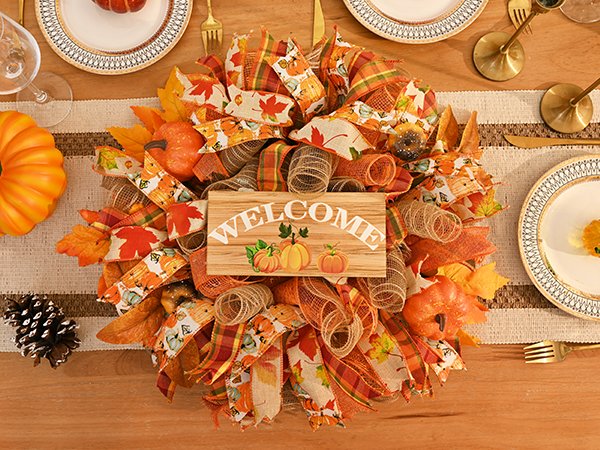 Fall mesh wreath with pumpkins, leaves, and a wooden 'Welcome' sign, set on a table with place settings.