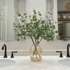 Faux eucalyptus stems in a glass vase on a bathroom vanity between two sinks.