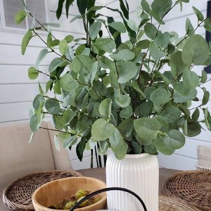Faux eucalyptus stems with green leaves in a white ribbed vase on a table with a wooden bowl.