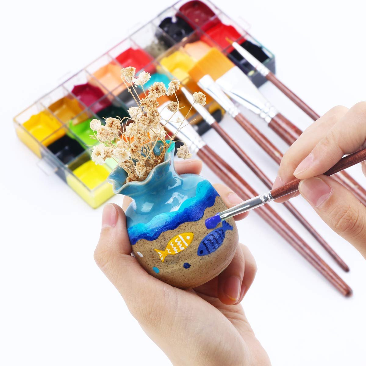 Hands painting a small ceramic vase with fish designs using a brush and paint palette.