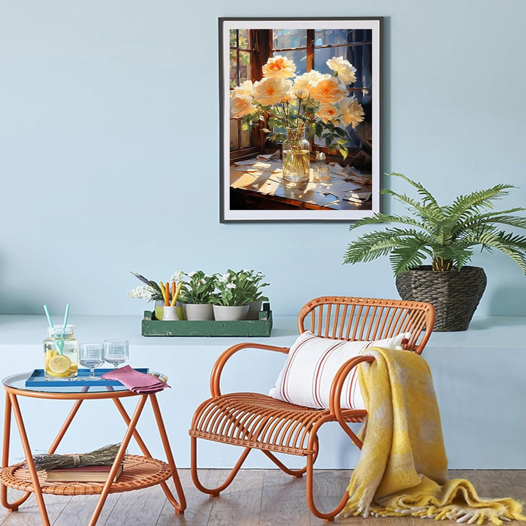 Framed painting of yellow roses in a vase on a windowsill with scattered papers, displayed in a room with a chair and plants.
