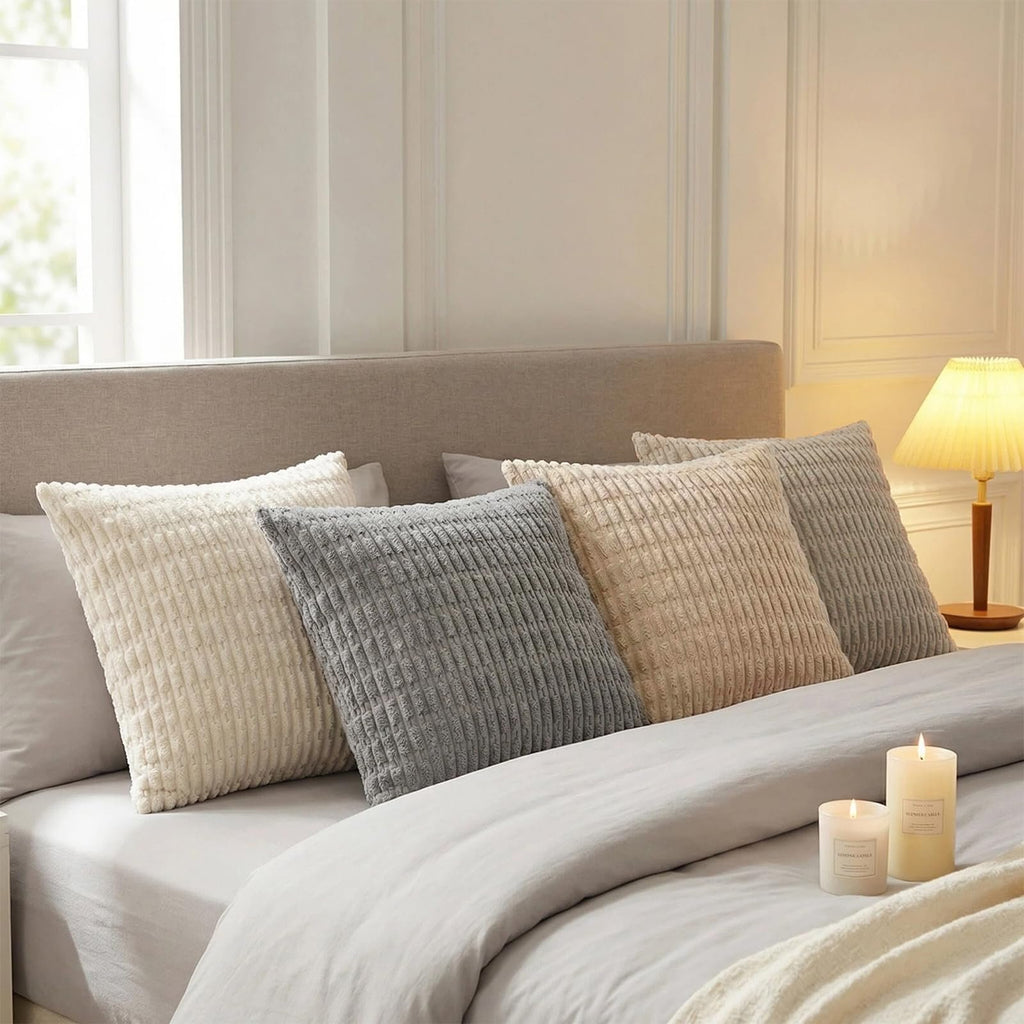 Four decorative throw pillow covers in neutral tones arranged on a bed with a headboard and lamp.