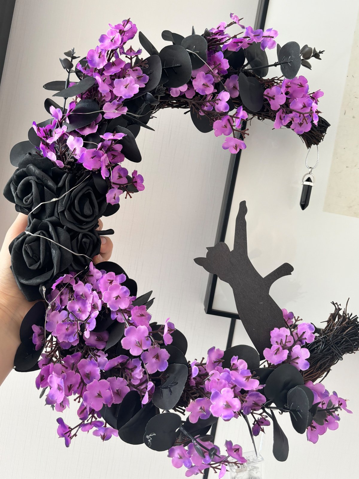 Halloween wreath with purple flowers, black roses, black cat silhouette, and hanging crystal charm held by a hand.