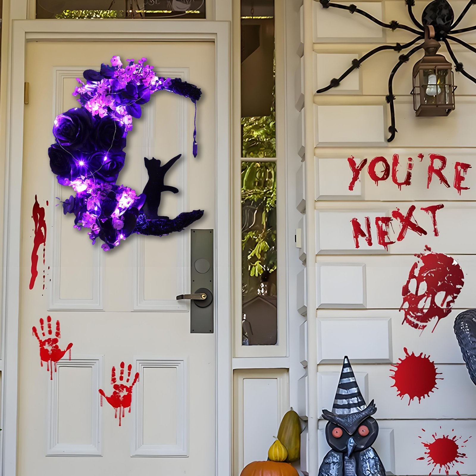 Halloween wreath with black cat on moon, purple flowers, and lights, hanging on a front door with bloody handprints and skull decorations.