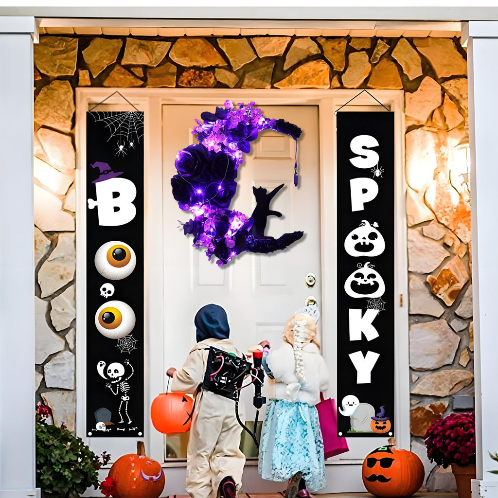 Halloween porch decor with a lighted crescent moon wreath and two vertical banners, one reading BOO and the other SPOOKY, with children in costumes approaching the door.
