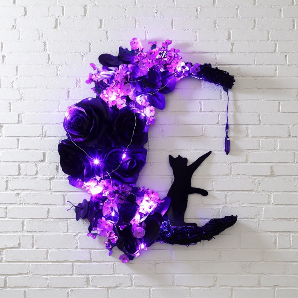 Halloween moon wreath with black cat, purple flowers, greenery, and purple lights on a white brick wall