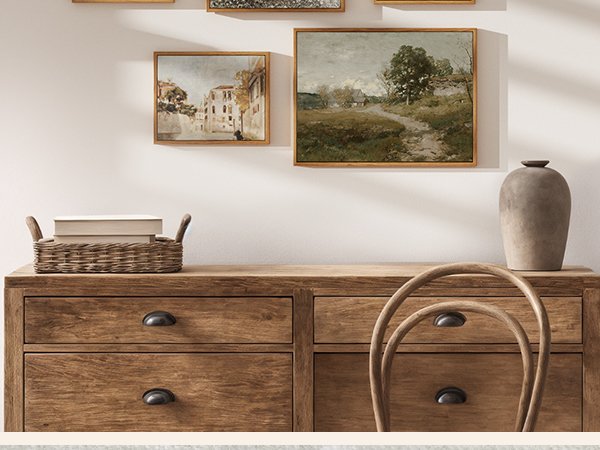 Two framed vintage landscape paintings displayed on a wall above a wooden dresser with a woven basket of books and a vase.