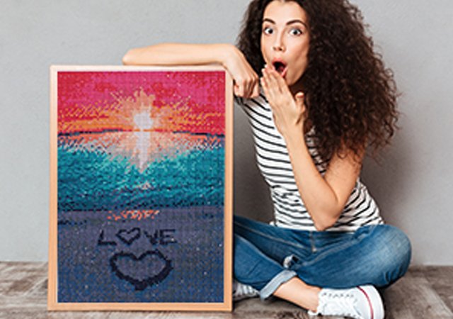 Woman with surprised expression holding a framed diamond painting kit of a beach sunset with the word LOVE written in the sand
