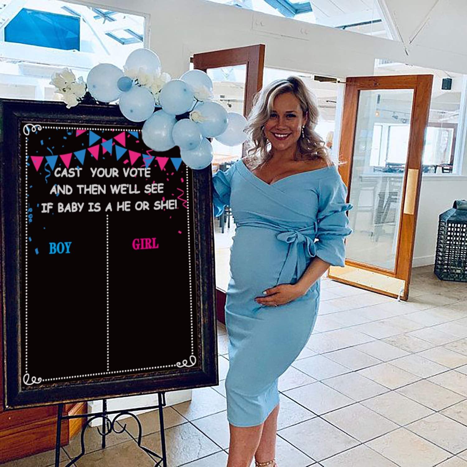 Pregnant woman stands next to a gender reveal voting board with 'Boy' and 'Girl' columns, decorated with balloons.