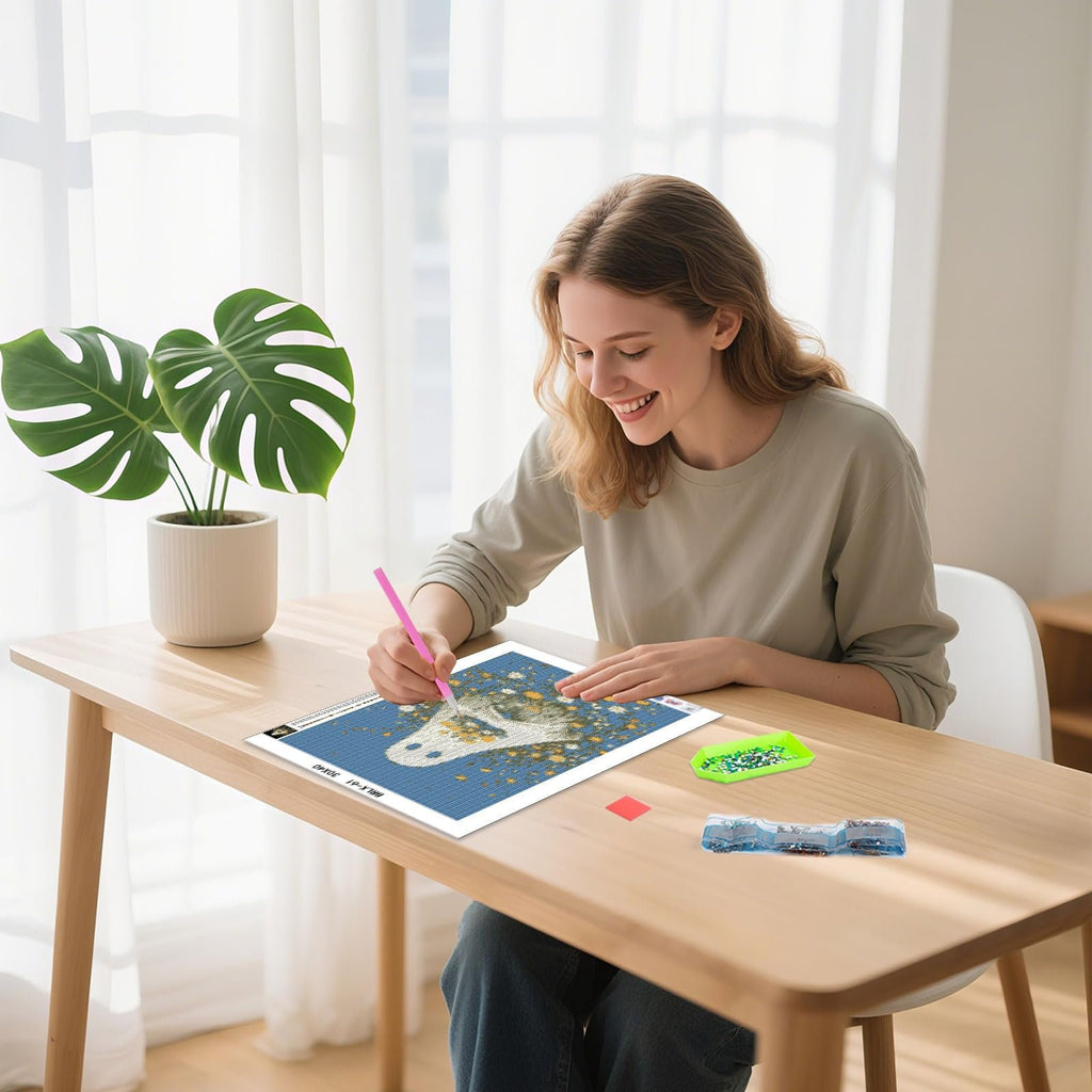Woman smiling while working on a diamond painting kit of a ghost flower on a wooden desk with plants in the background