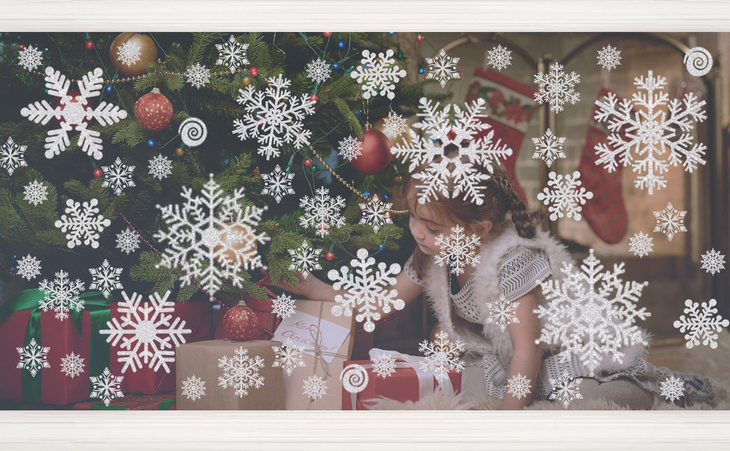 Glitter snowflake window clings applied to a window with a Christmas tree and gifts visible behind them.