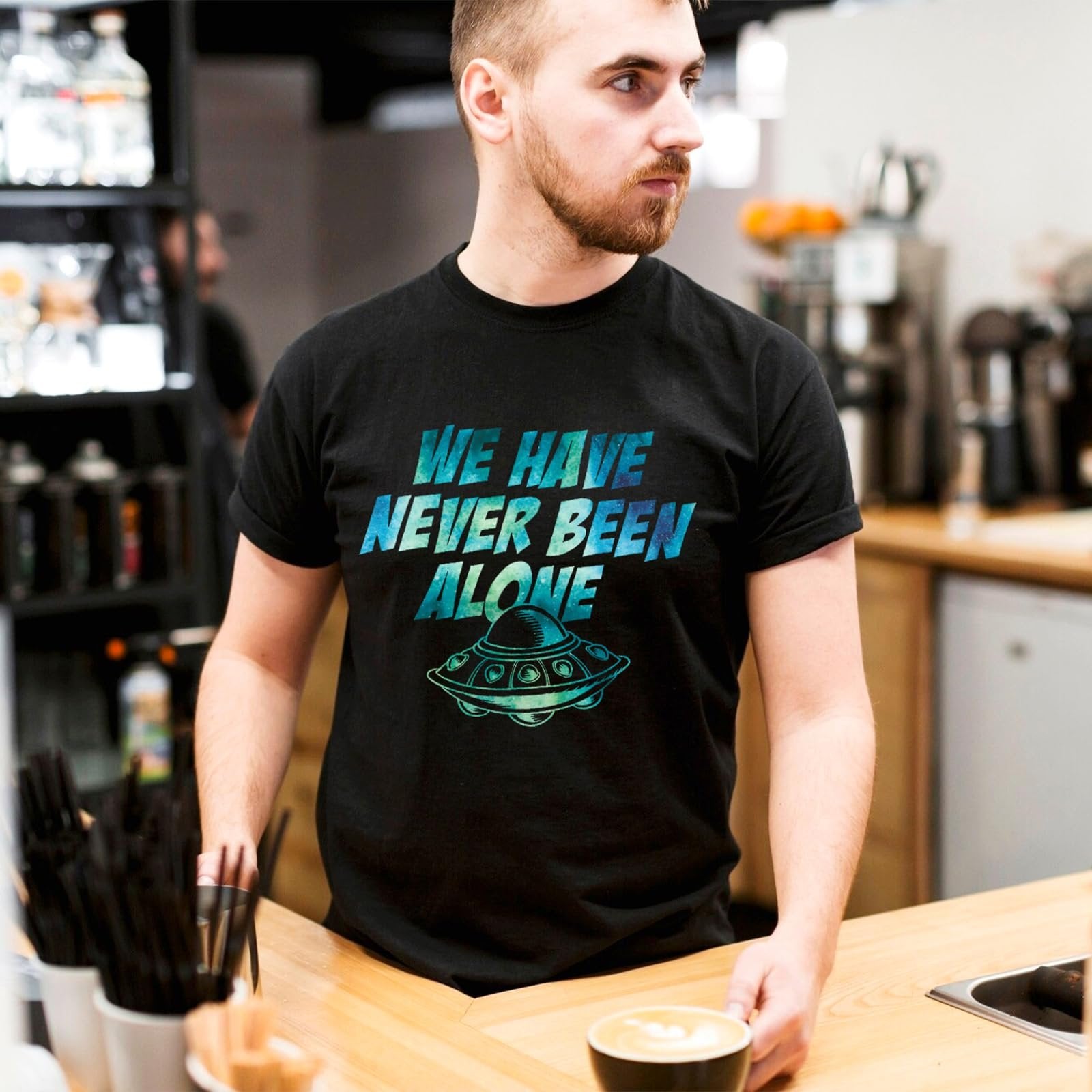 Man wearing a black t-shirt with a graphic of a UFO and the text "WE HAVE NEVER BEEN ALONE" in blue and green.