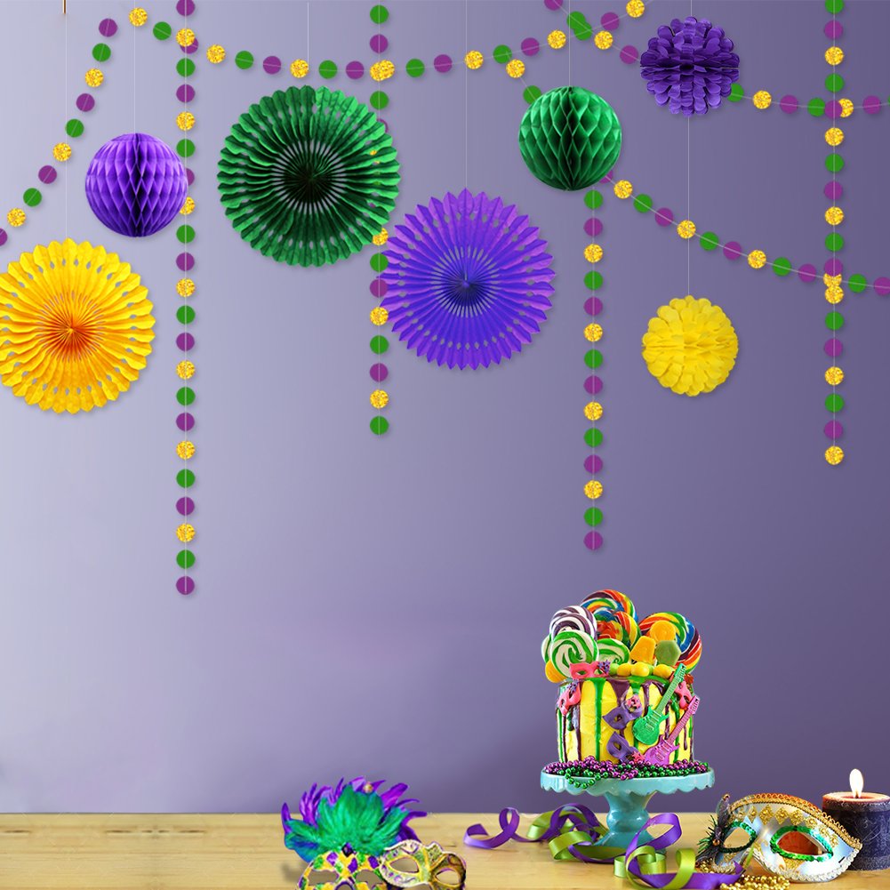 Mardi Gras themed party decorations including garlands, honeycomb balls, and paper fans displayed above a cake with lollipops and masks on a wooden surface.