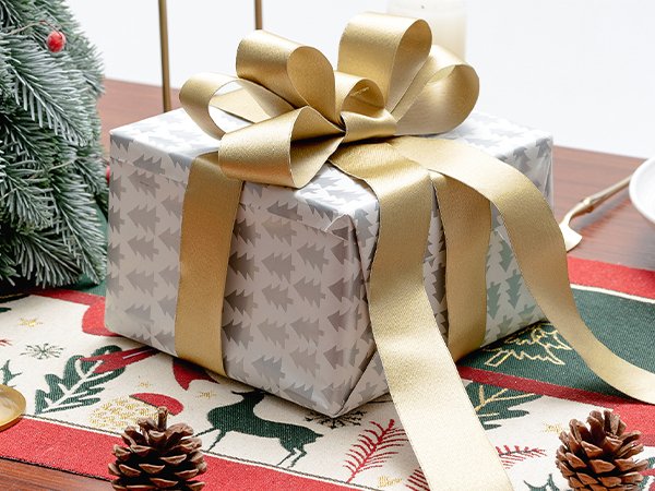 Gift-wrapped box with a gold satin ribbon bow, next to a frosted Christmas tree and pinecones.